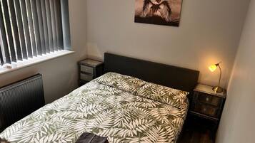 Un dormitorio con cama, mesita de noche, lámpara, radiador y ventana con persianas.