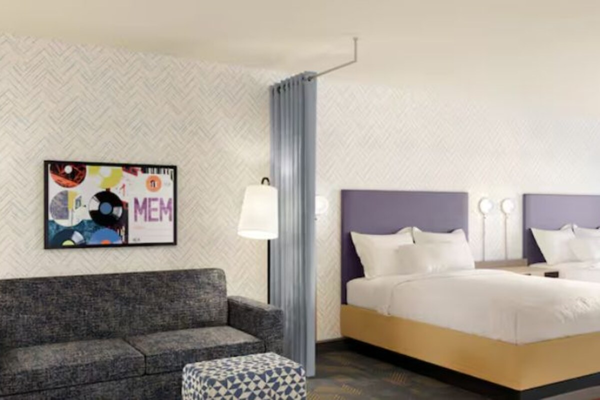 Una habitación de hotel con un sofá gris, una cama con ropa blanca y una obra de arte colgada en la pared.