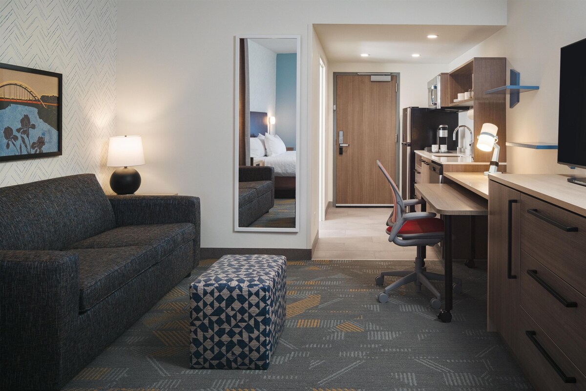 A hotel room with a sofa, ottoman, desk, chair, kitchenette, and a large mirror.