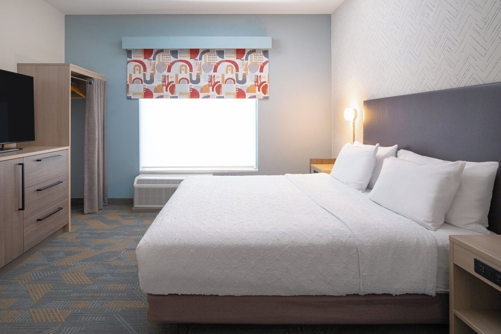 Een hotelkamer met een bed, een televisie, een garderobe, een raam met gordijnen en een wandlamp.
