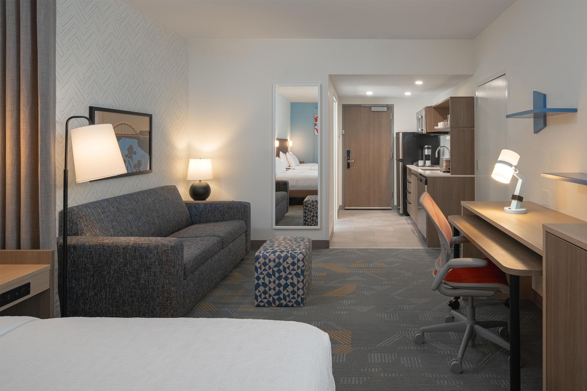 A hotel room with a bed, a sofa, a desk, a chair, a mirror, a lamp, and a kitchenette.