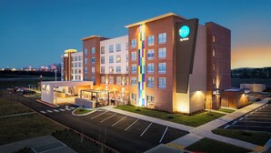 Un hotel de varios pisos con un llamativo letrero 'tru by Hilton', un estacionamiento y una zona ajardinada con un pequeño jardín.