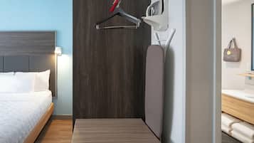 A hotel room with a bed, a small table, a wall-mounted ironing board, a towel rack, and a hanging bag.