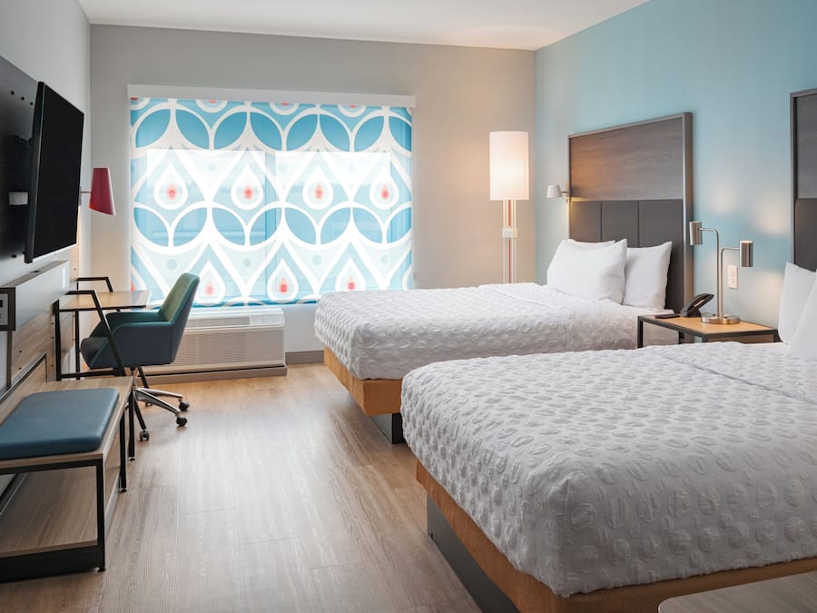 A hotel room with two beds, a desk, a chair, a TV, and a patterned window.