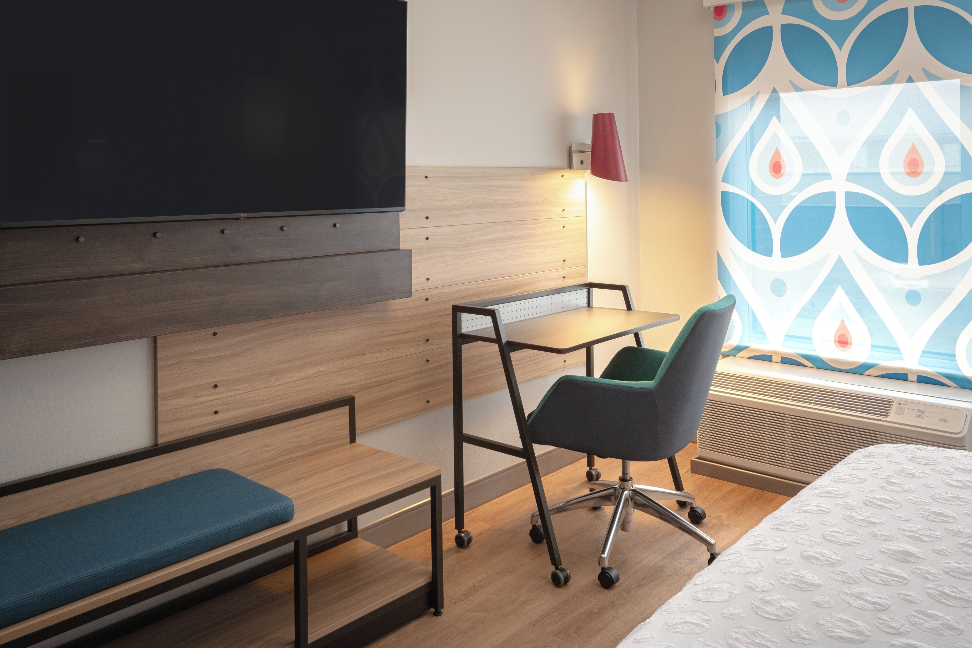 A hotel room with a bed, a desk, a chair, a bench, a wall-mounted TV, a heater, and a patterned curtain.