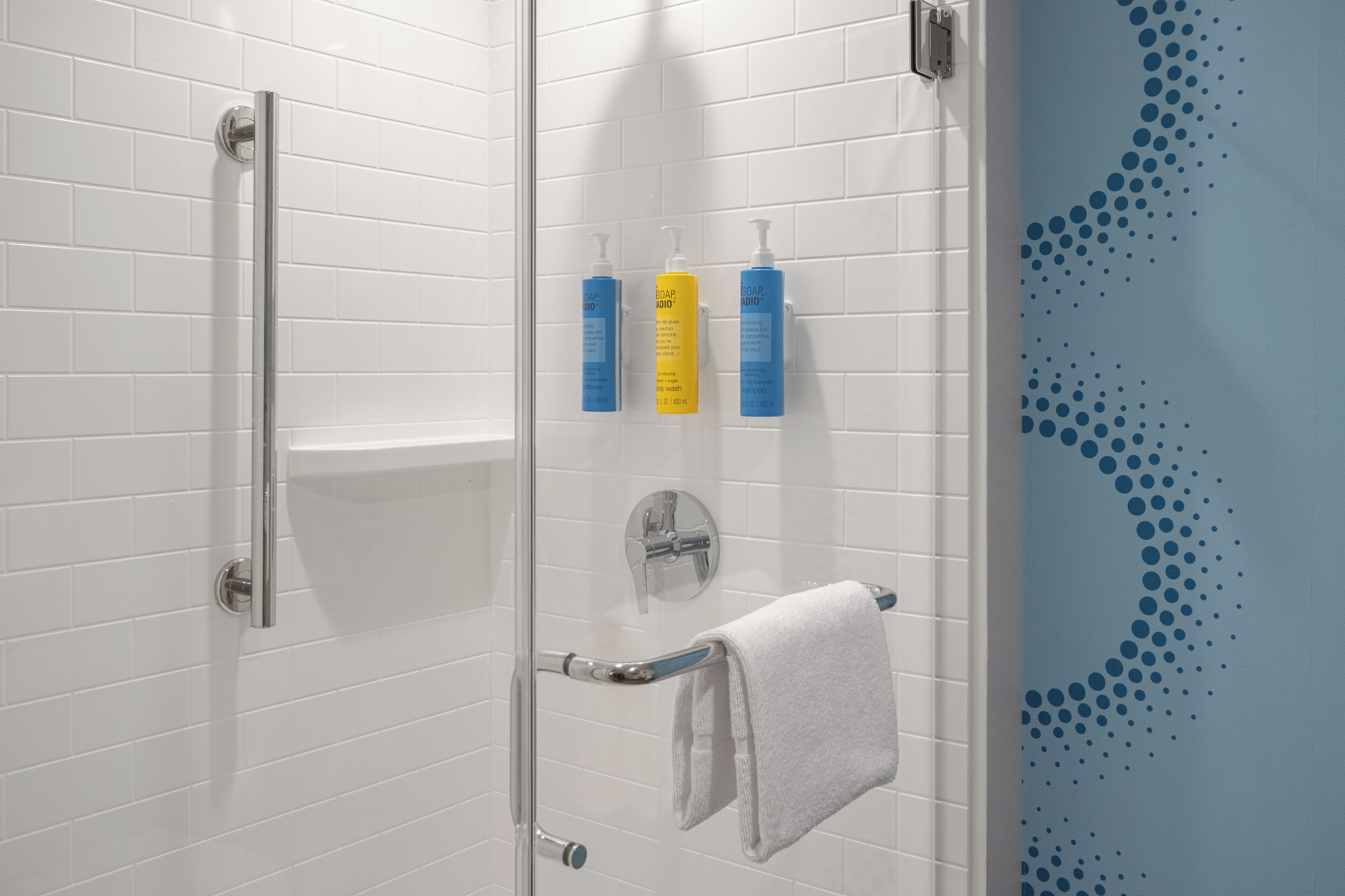 A shower with a glass door, a white towel hanging, and three soap dispensers on the wall.
