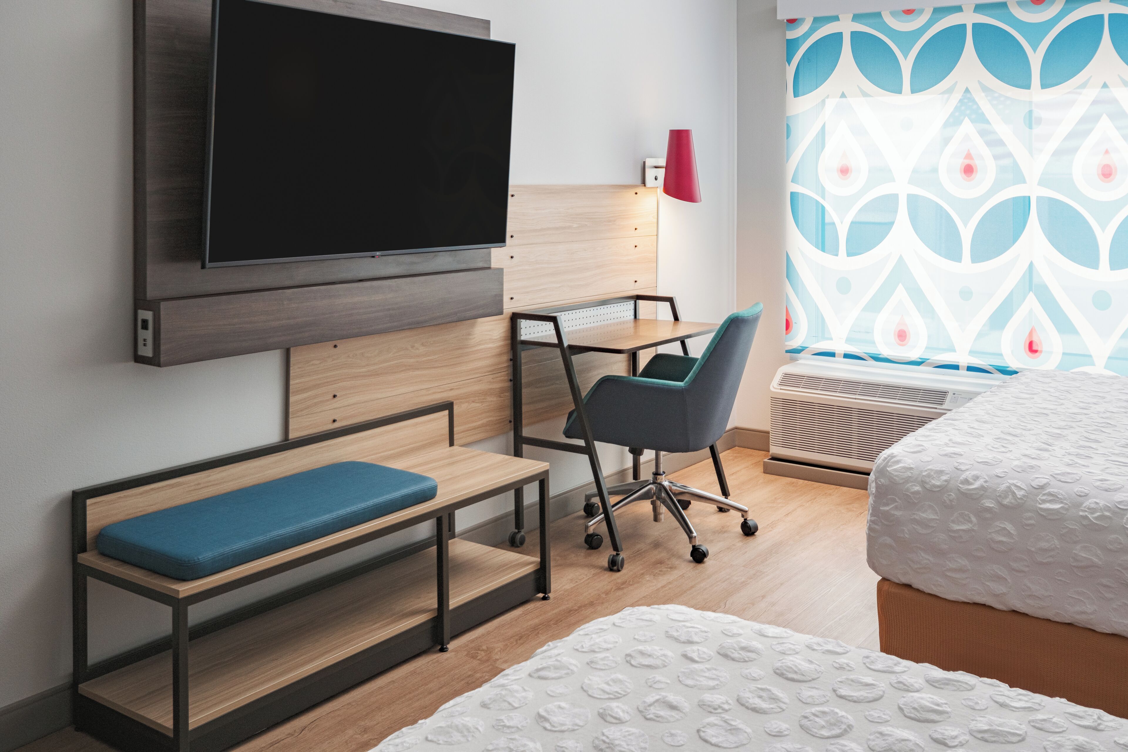 A hotel room with a bed, a desk with a chair, a television mounted on the wall, and a window with patterned curtains.