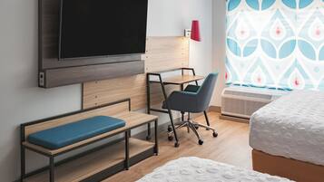 A hotel room with a bed, a desk with a chair, a television mounted on the wall, and a window with patterned curtains.