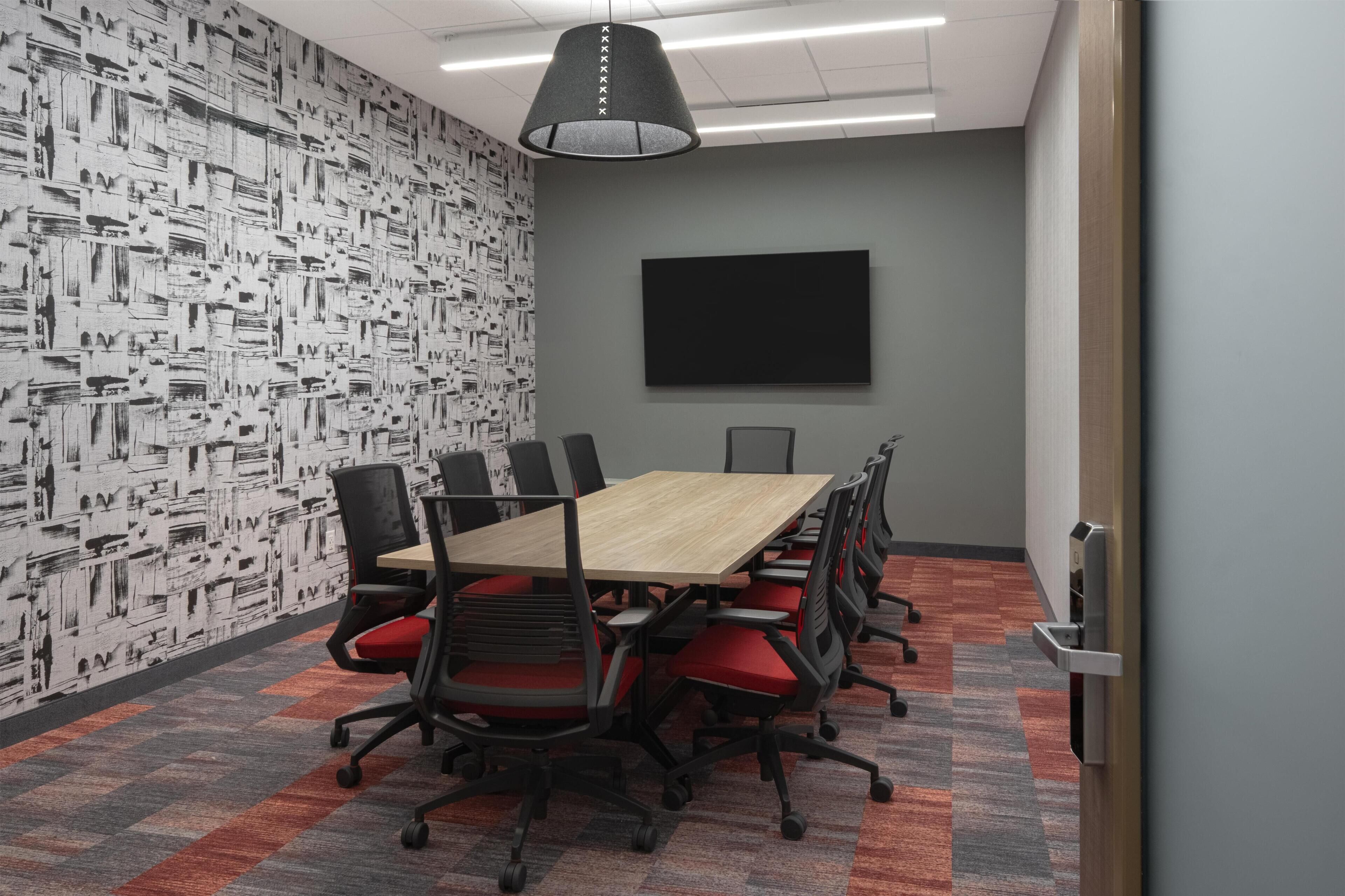 A conference room with a large table, red chairs, a black TV screen, and patterned wallpaper.