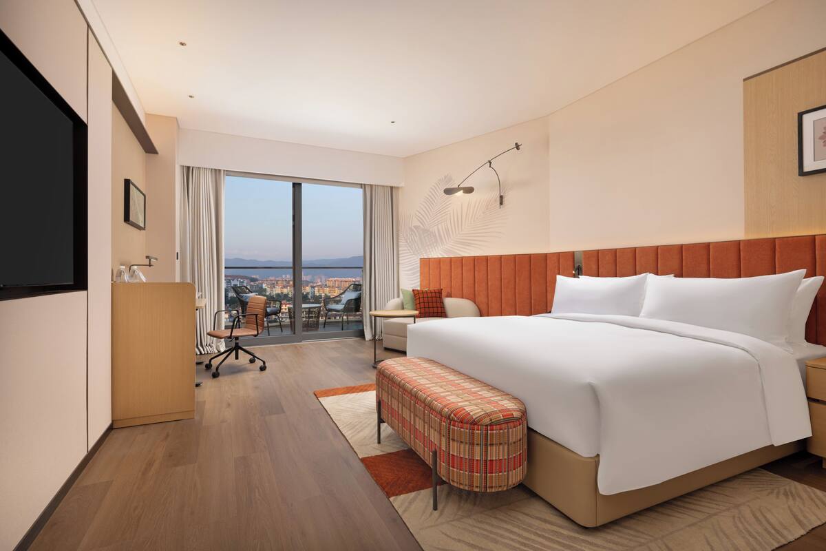 Habitación de hotel con una cama grande, un escritorio, una silla, un banco, un televisor y vistas de la ciudad.
