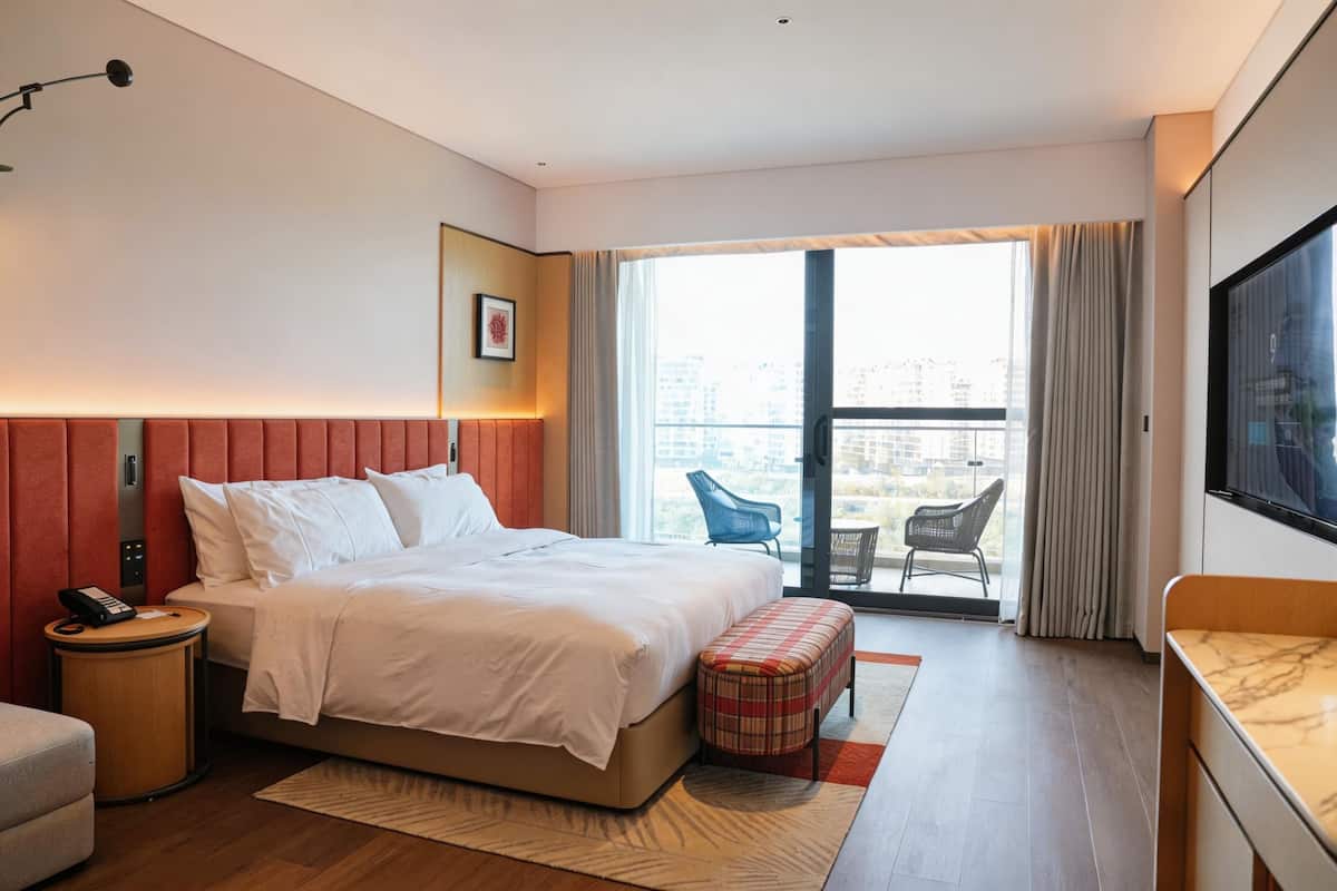 Habitación de hotel con cama, mesita de noche con teléfono, una pequeña mesa redonda, un banco, televisión y balcón con sillas.