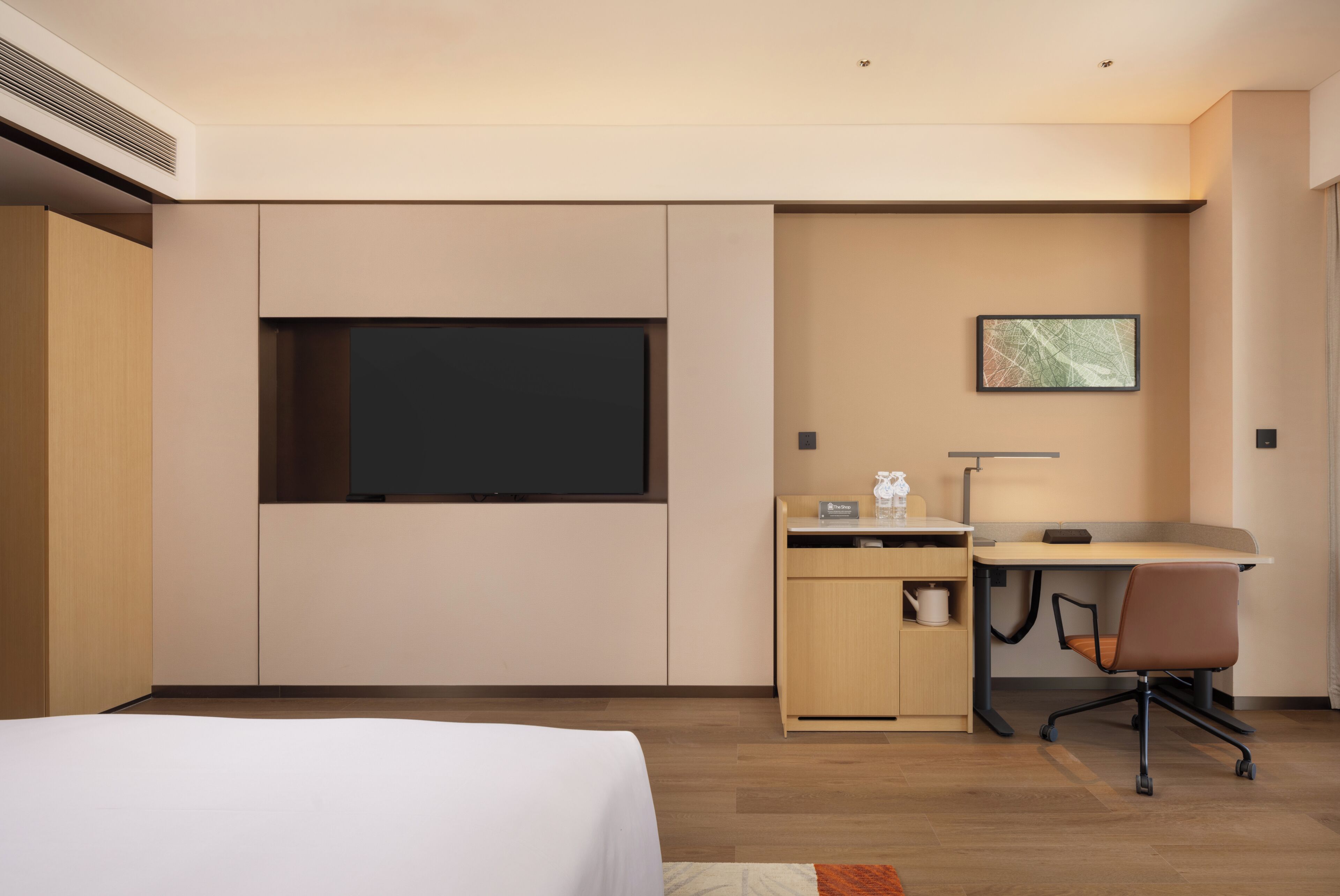 A modern hotel room with a bed, a desk with a chair, a flat-screen TV, a small cabinet, and a framed picture on the wall.