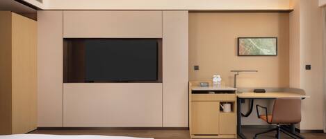A modern hotel room with a bed, a desk with a chair, a flat-screen TV, a small cabinet, and a framed picture on the wall.
