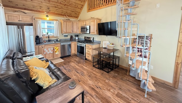 A cozy cabin-style kitchen with wooden cabinets, a stainless steel refrigerator, a microwave, and a stove. There's a dining area with a table and chairs, and a black leather couch with a yellow throw pillow.