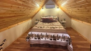 A cozy attic bedroom with a sloped wooden ceiling, a bed with a forest-themed bedspread, and two bedside lamps.