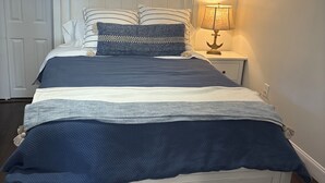 Ein Schlafzimmer mit einem weißen Bett, gestreiften Kissen, einer blauen Decke und einem Nachttisch mit einer Lampe.