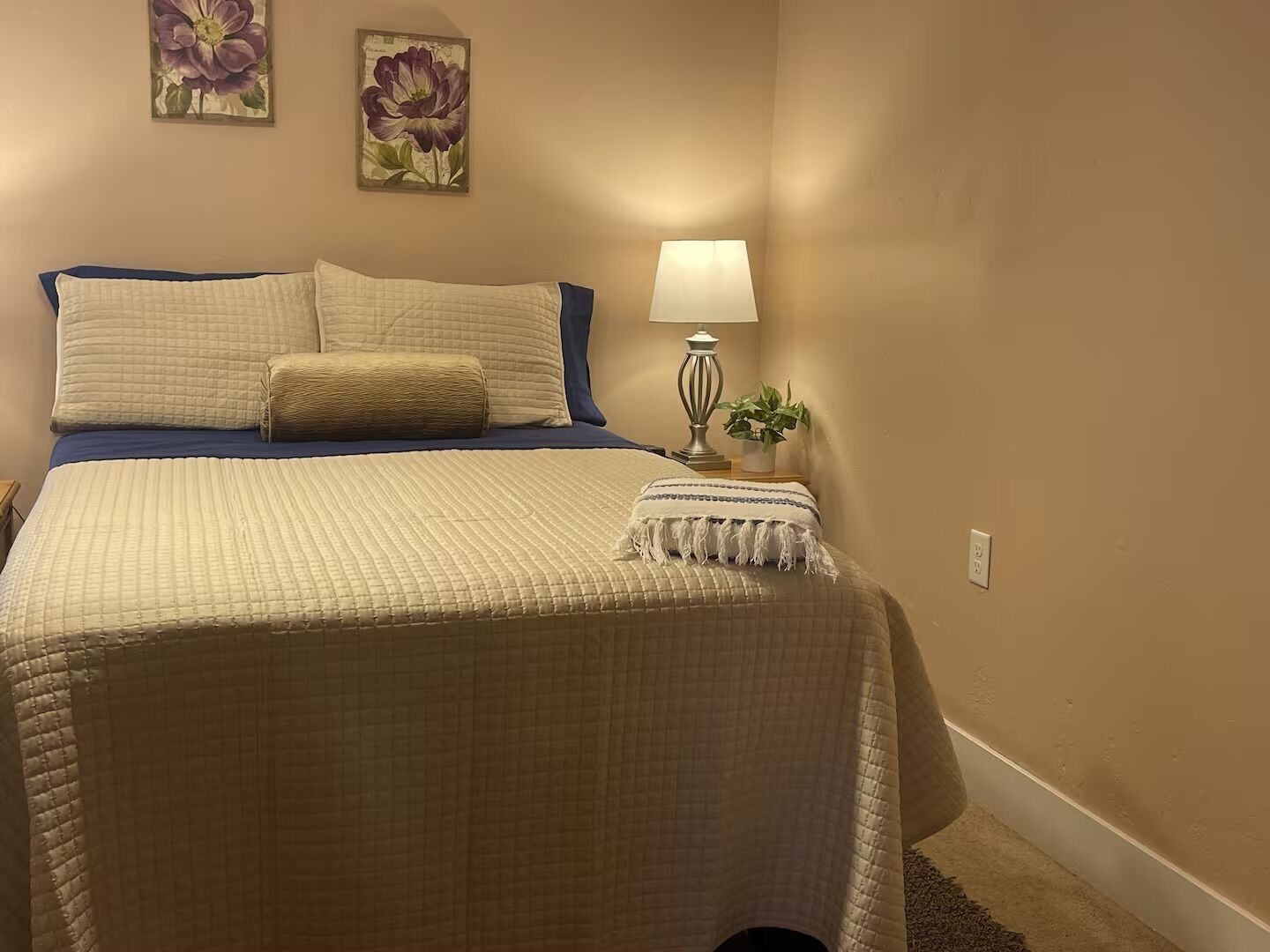 A bedroom with a bed, pillows, a bedside lamp, a small plant, and two floral wall art pieces.