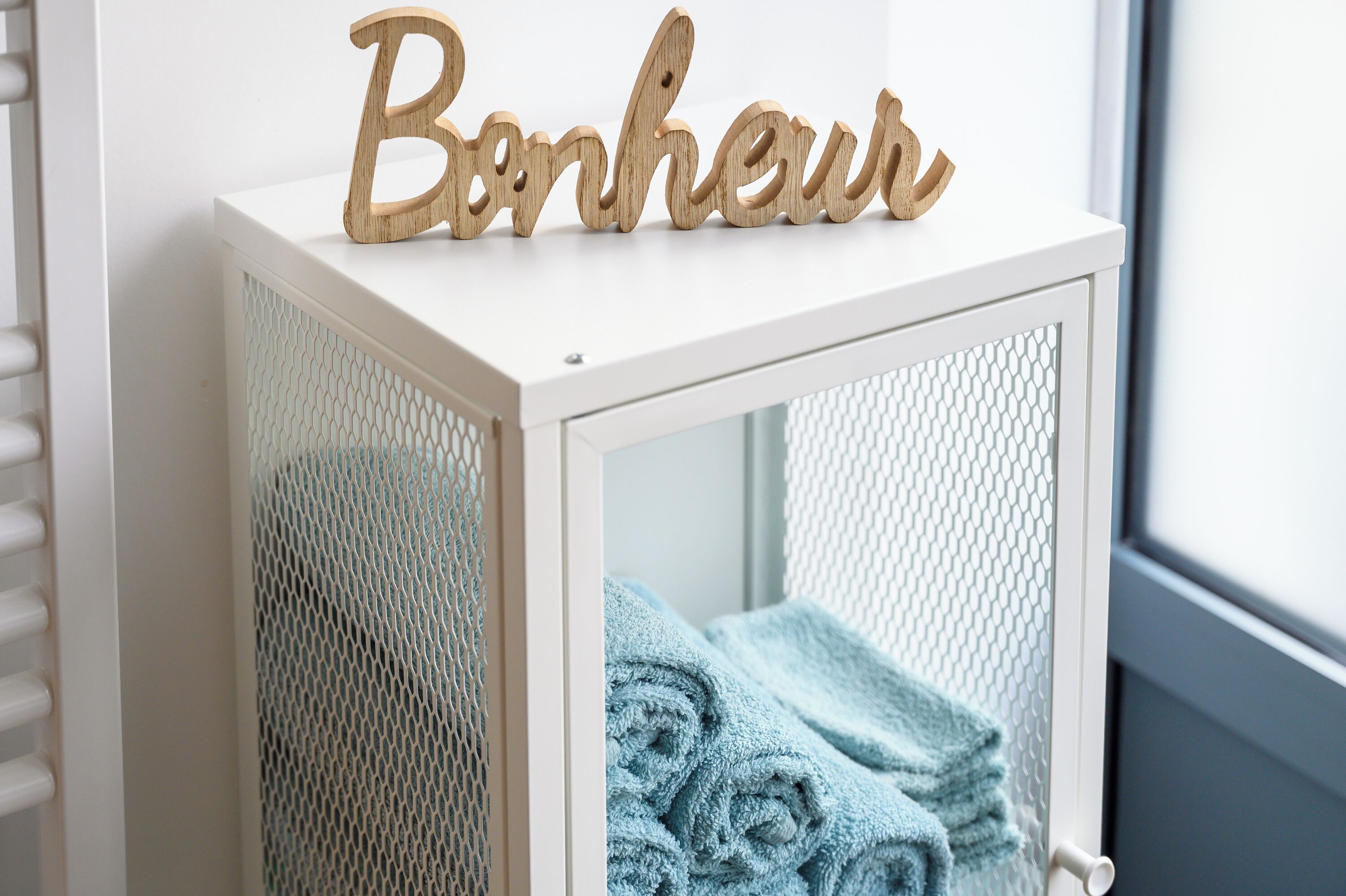 A white cabinet with a mesh door, a wooden sign spelling "Bonjour," and a stack of blue towels inside.
