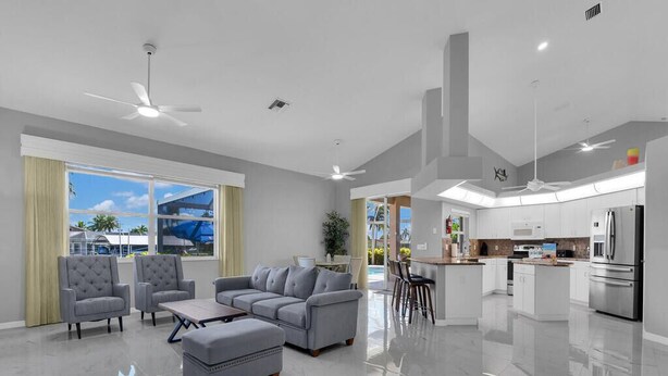 Uma sala de estar moderna com um conjunto de sofá cinza, uma mesa de centro e uma televisão. Há uma área de cozinha com armários brancos e uma geladeira de aço inoxidável.