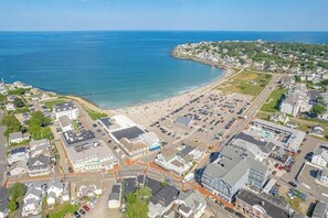 An aerial view of a coastal town with a beach, parking lots, and residential buildings.