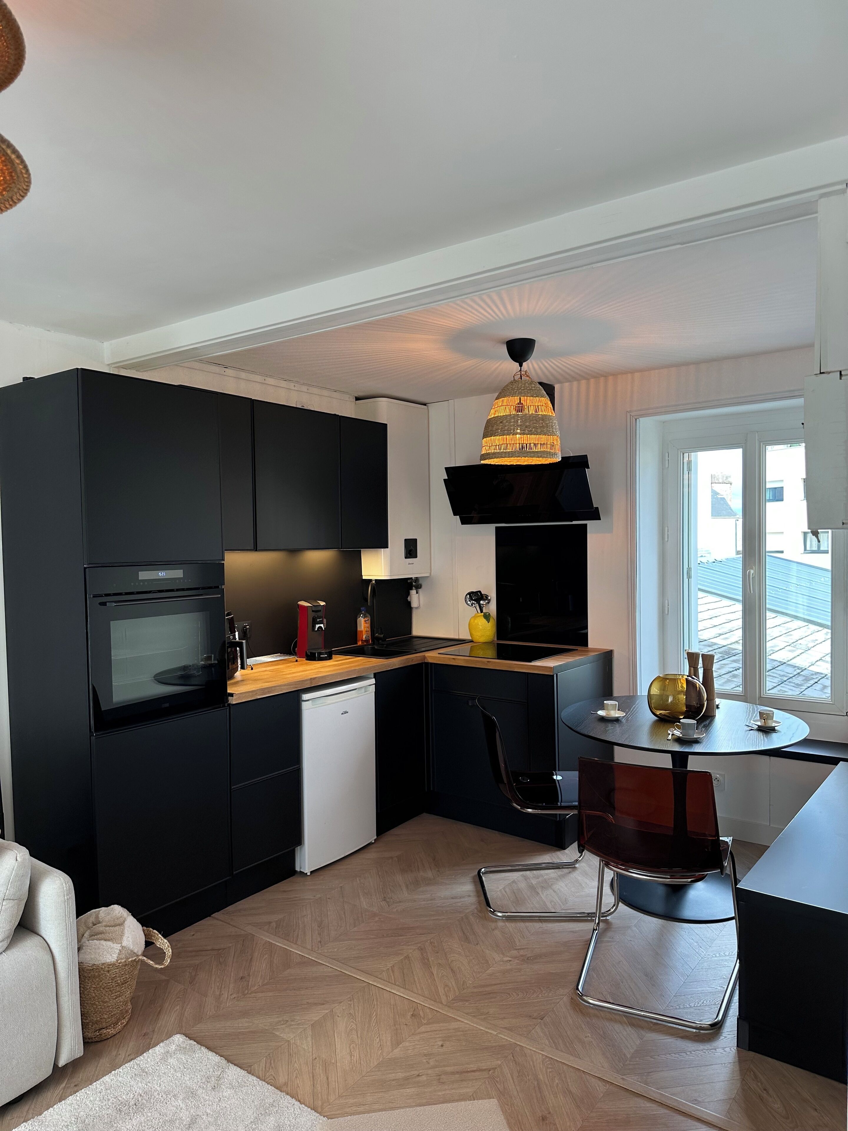 Une cuisine moderne avec des meubles de couleur noire, un four encastré et un petit lave-vaisselle. On y trouve un coin repas avec une table ronde et des chaises, ainsi qu’une grande fenêtre offrant une vue sur l’extérieur.