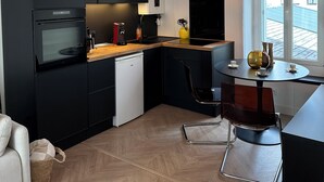 Una cocina moderna con armarios negros, horno empotrado y lavavajillas pequeño. Hay una zona de comedor con una mesa redonda y sillas, y un ventanal con vistas al exterior.