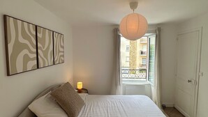 Un dormitorio con una cama, una mesita de noche, una lámpara de pared y una ventana con vistas a edificios.