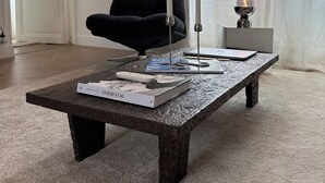 Un salón con una mesa de centro de madera oscura, un sillón negro, una mesita auxiliar con una lámpara y una puerta al fondo.