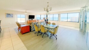 Een ruime woonkamer met een eettafel, stoelen en een rode kast. Er zijn grote ramen met uitzicht op de oceaan en gebouwen.