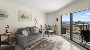 A living room with a grey sofa, a small table, and a painting of a coastal scene. There's a balcony with a view of the beach and palm trees.