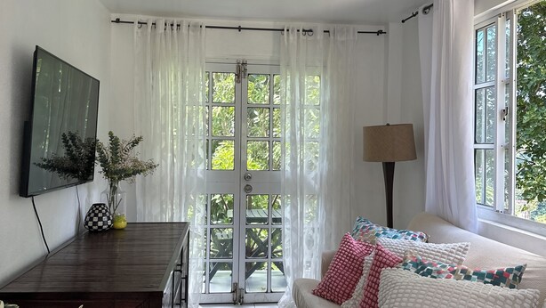 Ein Zimmer mit einer Couch, einem Schreibtisch und einem Fernseher. Es gibt Fenster mit Vorhängen und Blick ins Grüne.