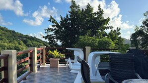 Eine Terrasse im Freien mit weißen Stühlen und Tischen, einem Holzzaun und Blick auf einen grünen Hügel.