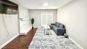 Un salon avec un canapé d’angle gris, une table basse blanche, un tapis moelleux, une cheminée blanche et une porte avec des volets blancs.
