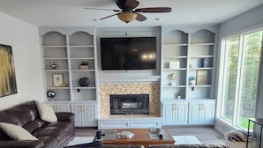 A living room with a fireplace, a large TV, and a ceiling fan. - Cutie On The Corner (Omaha)