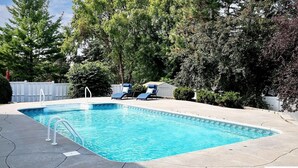 A rectangular swimming pool with a clear blue water, surrounded by a concrete deck. There are two lounge chairs with blue cushions and a white fence in the background. - Cutie On The Corner (Omaha)