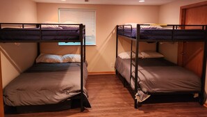 Two bunk beds with wooden frames and grey bedding, a window with blinds, and a wooden door. - Cutie On The Corner (Omaha)