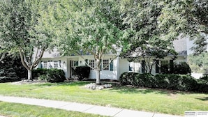 A two-story house with a front porch, surrounded by trees and a well-maintained lawn. - Cutie On The Corner (Omaha)