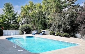 Una piscina rectangular con agua azul cristalina, rodeada por una plataforma de hormigón. Hay dos sillones de playa con cojines azules y una valla blanca al fondo.