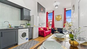 Une cuisine moderne équipée d’un lave-linge, un coin repas avec une table dressée pour deux personnes, et un salon avec un canapé et des coussins rouges.