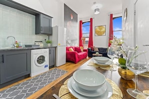 Une cuisine moderne équipée d’un lave-linge, un coin repas avec une table dressée pour deux personnes, et un salon avec un canapé et des coussins rouges.