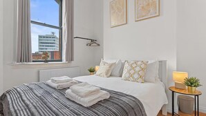 Ein Schlafzimmer mit einem großen Bett, einem fensterbelaubten Fenster, einem Heizkörper, einem Nachttisch mit Lampe und einer Wandleuchte.
