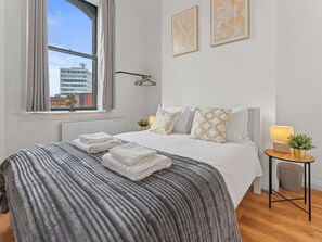 Un dormitorio con una cama grande, una ventana con cortinas, un radiador, una mesita de noche con lámpara y una luz empotrada en la pared.