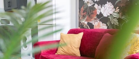Um sofá vermelho com um travesseiro bege em um quarto com máquina de lavar e um vaso de plantas.
