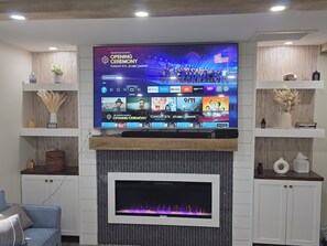 Uma sala de estar com uma televisão de tela plana sobre uma lareira de madeira, exibindo a cerimônia de abertura dos Jogos Olímpicos de Inverno.