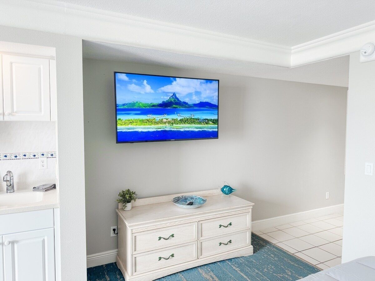 A room with a flat-screen TV mounted on the wall, a white dresser with drawers, a small plant, and a decorative fish bowl.