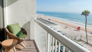 Varanda com cadeira de vime e mesa pequena, com vista para uma praia com torre salva-vidas e palmeiras.