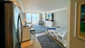 A compact hotel room with a kitchenette, a bed with a patterned bedspread, a desk with a chair, a large window with a view of the sea, and a ceiling fan. - The Reserve | Oceanfront Studio | Private Balcony (Daytona Beach)