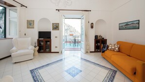 Uma sala de estar com um sofá branco, uma poltrona branca, uma televisão e um espaço de jantar com mesa e cadeiras.