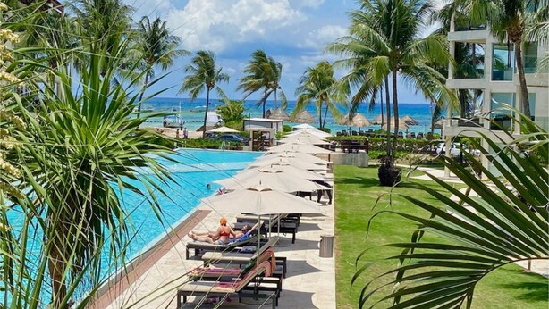 A resort pool area with lounge chairs and umbrellas, surrounded by palm trees and a view of the ocean.