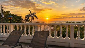 A balcony with two sun loungers, a lamp post, and a view of the sunset over the ocean.